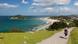 A hiker carries a baby on her back along a nature trail with the ocean and a town in the background